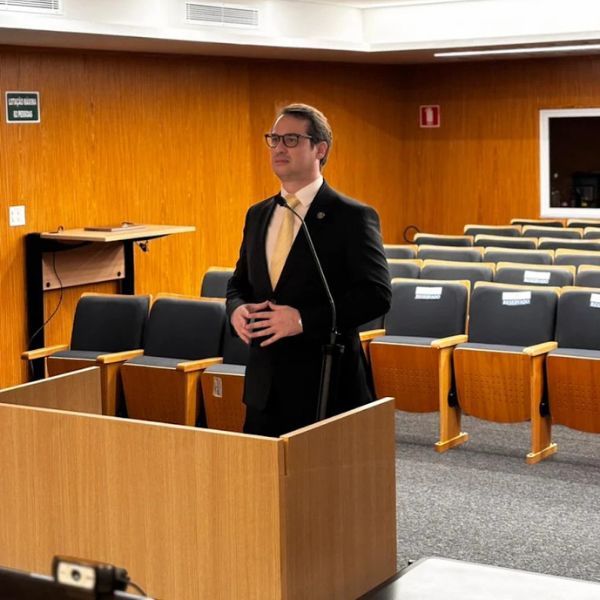 Filipe Augusto de Moura Meireles, advogado trabalhista, em traje social formal, terno escuro e gravata amarela, discursando ao microfone em auditório institucional com painéis de madeira, ambiente sério e elegante, fotografia realista