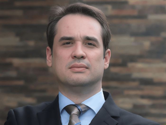 Retrato profissional de Filipe Augusto de Moura Meireles, advogado sócio trabalhista, homem jovem em traje social formal, terno escuro e gravata, postura séria e confiante, fundo neutro cinza-claro, iluminação de estúdio suave, fotografia corporativa realista