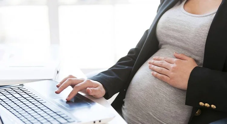 Imagem de uma mulher branca grávida com a mão na barriga em frente ao notebook pesquisando sobre direitos trabalhistas da gestante.